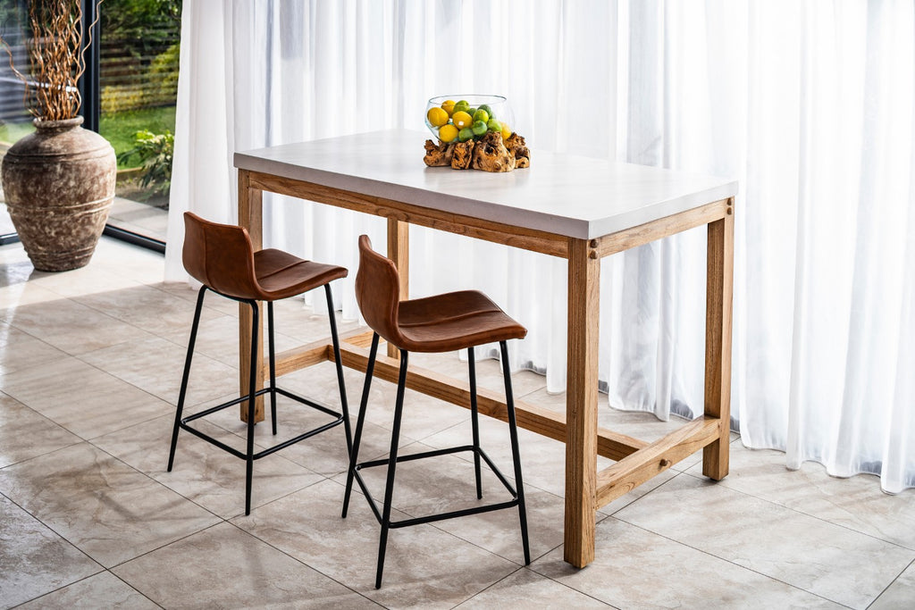 Wooden bar table with two stools in a bright room with white curtains.